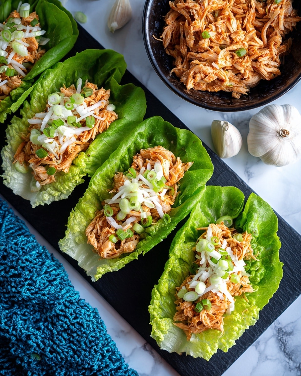 The image shows four bright green lettuce cups arranged in a row on a black board. Each cup holds a layer of finely chopped cooked chicken that is light brown with some pieces showing a slight shine from sauce, topped with thin white shredded vegetables and small pieces of green onions. Above them, there is a black bowl filled with more chopped chicken, and next to the bowl are three bulbs of white garlic. The background is a white marbled surface, with a blue textured cloth partially visible in the bottom left corner. Photo taken with an iphone --ar 4:5 --v 7