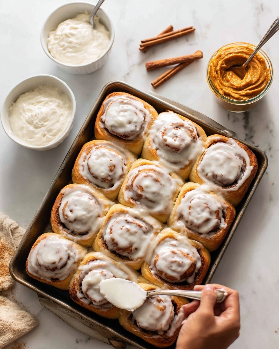 The image shows a baking tray filled with twelve cinnamon rolls that have a golden brown color and a thick layer of white icing spread unevenly on top of each roll, creating a slightly glossy texture. In the background on a white marbled surface, there are two small white bowls, one filled with more icing with a spoon inside, and the other with a pumpkin-colored spread and two cinnamon sticks placed diagonally on top. A woman’s hand is seen holding a small spoon dipped in the icing, about to spread it on the rolls. The scene is bright and warm, suggesting fresh baking. Photo taken with an iphone --ar 4:5 --v 7