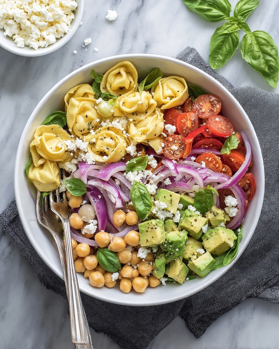 A white bowl filled with a colorful salad that has three main layers: the bottom layer is pale yellow tortellini pasta, the middle layer contains bright green avocado chunks and light pink chickpeas, and the top layer is made up of red cherry tomato halves, thin slices of purple onion, crumbled white cheese, and fresh green basil leaves as garnish. The bowl sits on a soft dark gray cloth placed on a white marbled surface. Two silver forks rest inside the bowl, leaning to the left. In the upper left corner, there is a white bowl filled with more crumbled cheese, and scattered green basil leaves are placed around the scene. photo taken with an iphone --ar 4:5 --v 7