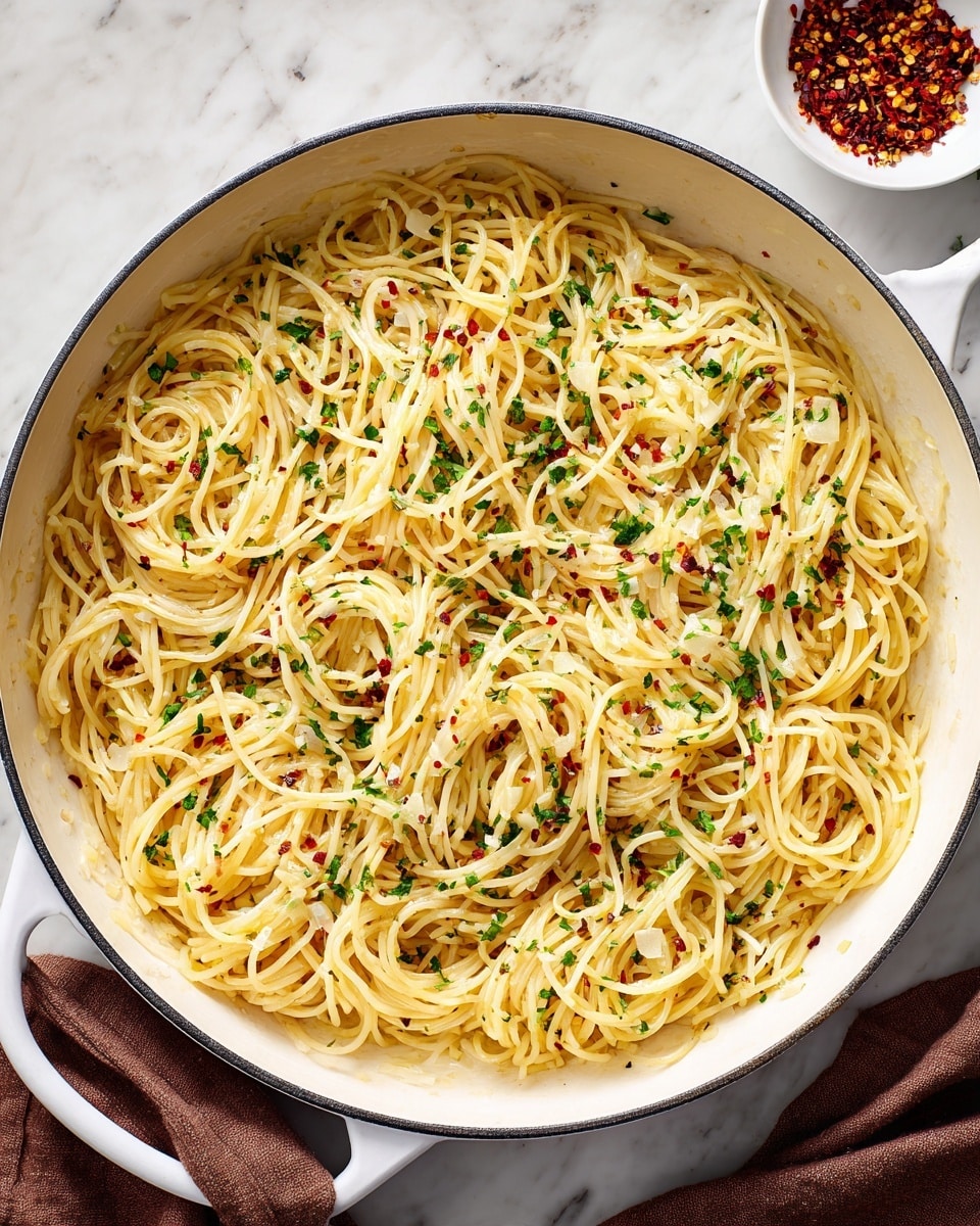 A white pan filled with a large amount of cooked spaghetti noodles mixed with thin slices of light white garlic scattered evenly throughout. The pasta has a light golden color with slight glints from oil, and small green herb bits are spread over it. Tiny red pepper flakes are sprinkled on top, adding small pops of red against the noodles. The pan rests on a white marbled surface with a small white bowl containing more red pepper flakes partly visible in the upper right corner and a brown cloth napkin in the lower right corner. Photo taken with an iphone --ar 4:5 --v 7