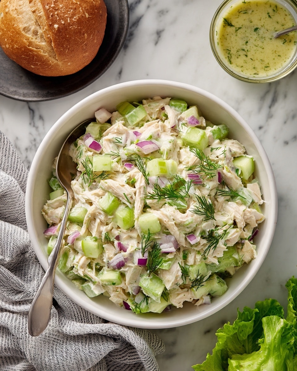 A white bowl filled with a layered salad made of shredded white chicken, chopped pale green pickles, diced light green celery, small bits of red onion, and sprinkled fresh green dill on top. A silver spoon rests inside the bowl on the left side. The bowl sits on a white marbled surface with a gray striped cloth partially visible on the left, a round bread roll in a black dish to the top left, a glass bowl of light yellow-green dressing with dill to the top right, and some green leafy lettuce at the bottom right. Photo taken with an iphone --ar 4:5 --v 7