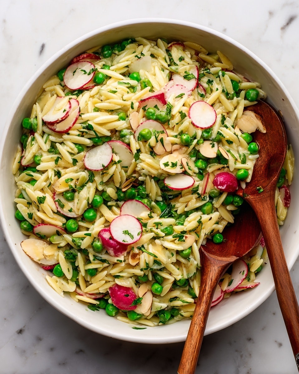 This image shows a white bowl filled with orzo pasta salad mixed with bright green peas and thin slices of radish with red edges and white centers. The salad is sprinkled with finely chopped green herbs and small pieces of almond. Two wooden spoons with light brown handles rest inside the bowl, slightly lifting the salad. The bowl is placed on a white marbled surface. The colors are fresh and vibrant, with the creamy yellow pasta, green peas, red and white radish slices, and scattered herbs creating a fresh look. photo taken with an iphone --ar 4:5 --v 7
