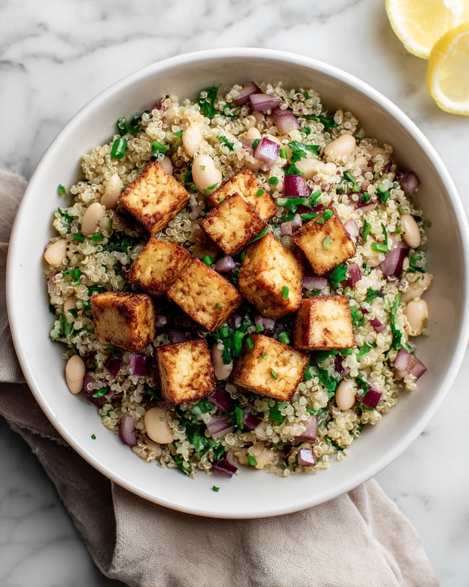 A white bowl filled with a quinoa salad that has about three visible layers: at the bottom, fluffy light beige quinoa with a slightly grainy texture; mixed throughout are small white beans and chopped green herbs, adding specks of green and cream. On top, golden brown crispy tofu cubes are evenly spread, mixed with bits of finely chopped purple onion and small green chive pieces. The bowl is set on a white marbled surface, with a lemon wedge on the right edge and a beige cloth napkin partially under the bowl. Photo taken with an iphone --ar 4:5 --v 7