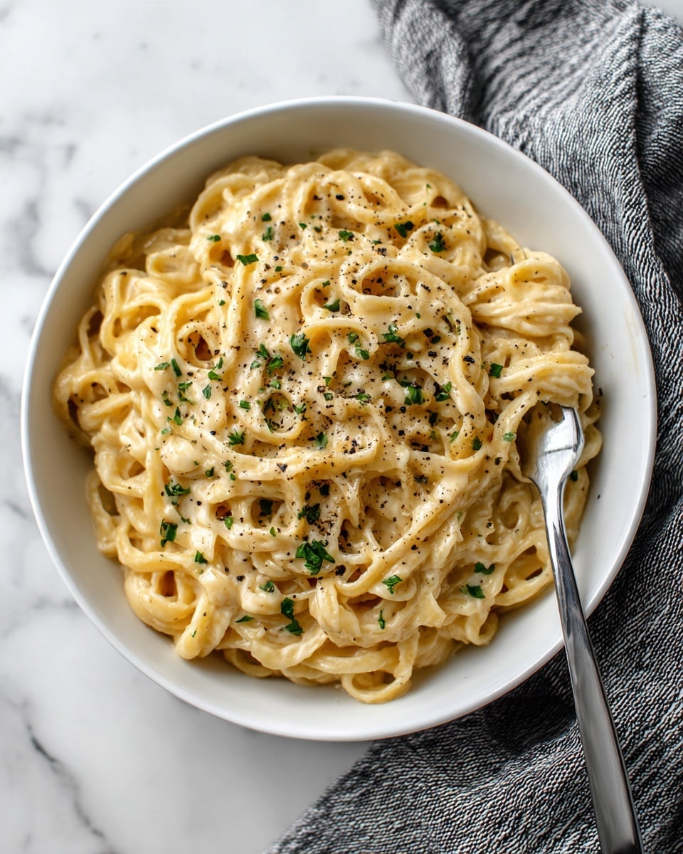 A white bowl filled with creamy pasta noodles coated in a light beige sauce, topped with small bits of green herbs and sprinkled with black pepper. The noodles are thick and curly, layered loosely and mixed evenly with the sauce. On the right side of the bowl, a fork picks up a small bundle of noodles. The bowl sits on a white marbled surface with a gray and white striped cloth nearby. Photo taken with an iphone --ar 4:5 --v 7