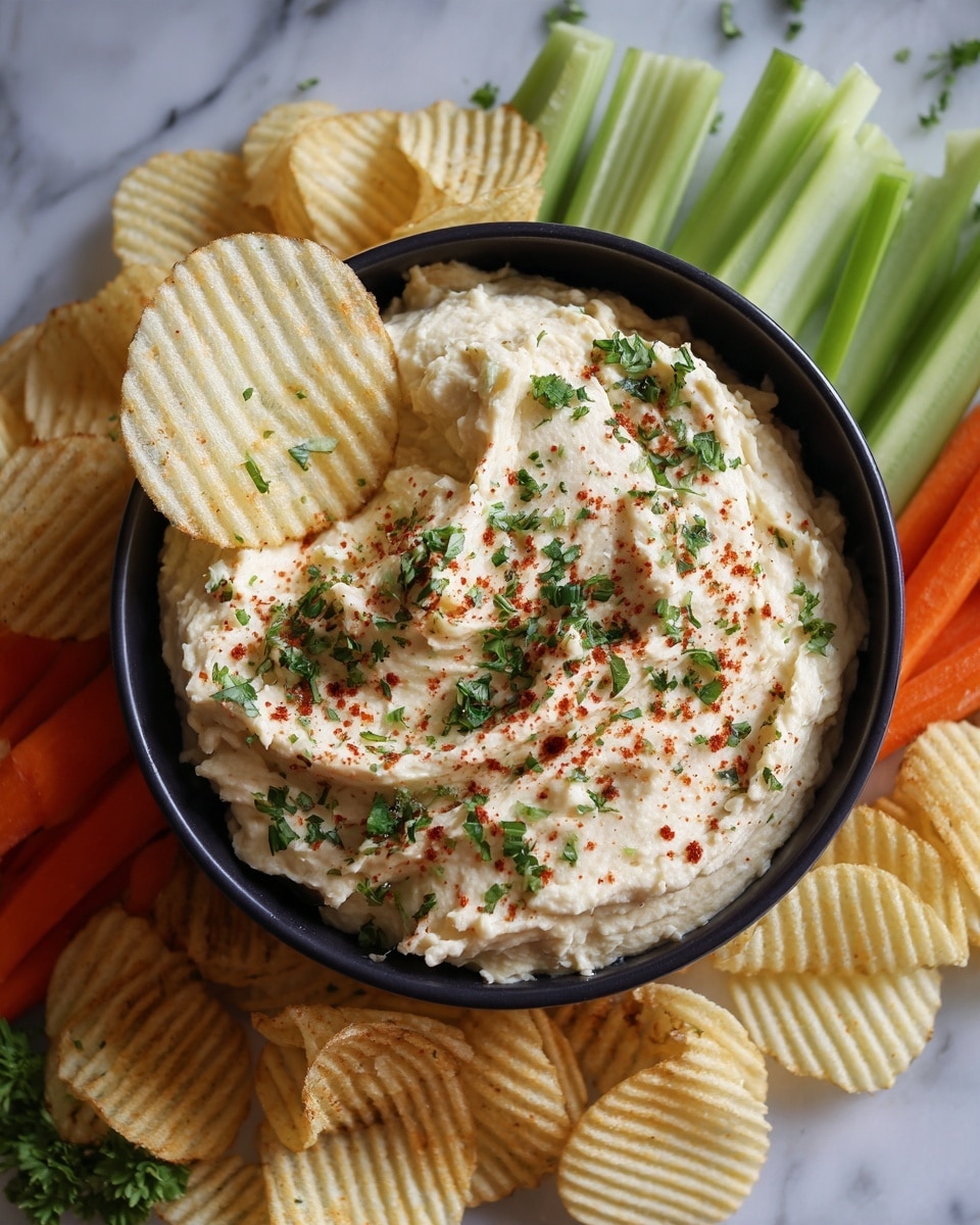 A black bowl filled with a creamy light beige dip sprinkled with finely chopped green herbs and a light dusting of red spice. A ridged potato chip, pale yellow with slight shine, is scooping up some dip from the bowl. Around the bowl, there is a border of ridged potato chips and fresh vegetables, including green celery sticks and orange carrot sticks, all placed on a white marbled surface. photo taken with an iphone --ar 4:5 --v 7