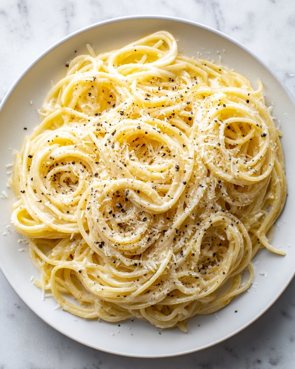 A close-up view of a white plate filled with creamy spaghetti, showing multiple long, smooth, and lightly yellow pasta strands tangled together. The pasta is coated in a pale yellow cream sauce with small bits of shredded cheese and black pepper sprinkled generously on top, creating small black and white specks over the creamy texture. The background is a white marbled surface. photo taken with an iphone --ar 4:5 --v 7
