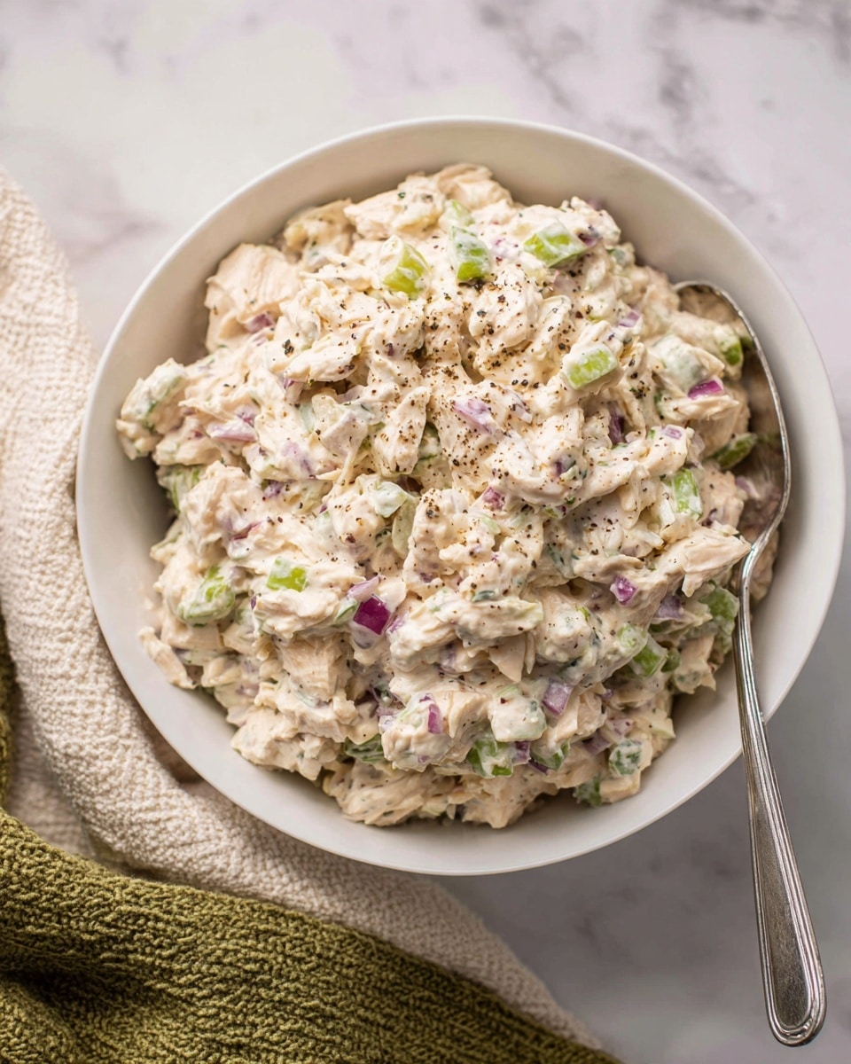 A close-up of a white bowl filled with three layers of creamy chicken salad mixed with small pieces of green celery and bits of purple onion scattered throughout. The creamy chicken salad is pale cream in color with rough, chunky texture. Light black pepper sprinkled on top creates tiny dark specks across the salad. A silver spoon rests inside the bowl on the right edge, extending outwards. The bowl sits on a white marbled surface with a cream textured cloth and a green knitted cloth near the bottom left corner. Photo taken with an iphone --ar 4:5 --v 7