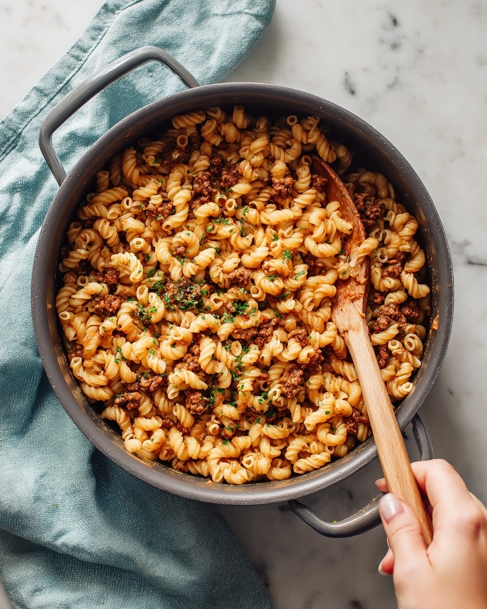 A dark gray pot filled with cooked curly macaroni pasta mixed with small chunks of browned meat and green herbs, giving it a warm reddish-brown color. A woman's hand holds a wooden spoon resting inside the pot, stirring the pasta. The pot is placed on a white marbled surface with a light blue cloth nearby. Photo taken with an iphone --ar 4:5 --v 7
