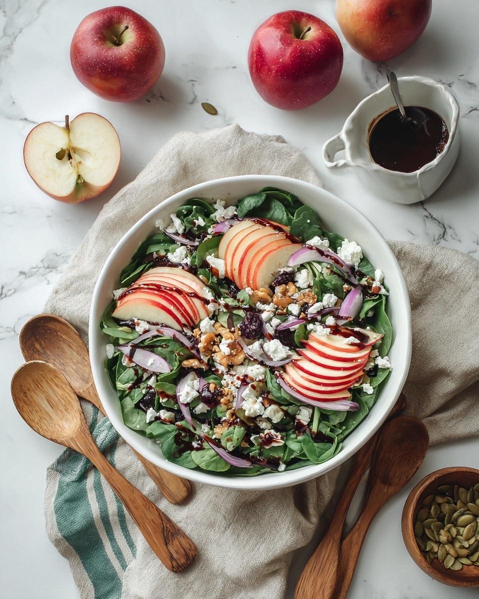 A white bowl filled with fresh green leafy spinach and arugula forming the base layer, topped with thin, bright red apple slices arranged in small overlapping groups on three sides, scattered bits of white cheese crumbles, small dark dried berries, and thin, curved slices of red onion adding purple hues across the salad. There are small pieces of chopped nuts and pumpkin seeds sprinkled unevenly on top, all drizzled with a thick dark balsamic dressing. The bowl sits on a beige cloth with a white and green striped towel underneath, on a white marbled surface. Nearby are two whole red apples, a few apple slices, a small white bowl filled with dark dressing with a spoon, and a wooden bowl with pumpkin seeds. Two wooden utensils lay next to the bowl. Photo taken with an iphone --ar 4:5 --v 7