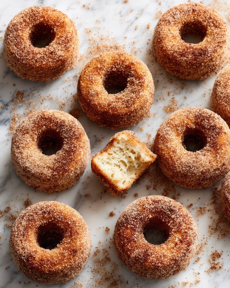 The image shows eight round donuts with a hole in the middle, all covered with a thick layer of brown cinnamon sugar. The donuts have a rough texture and a golden brown color underneath the sugar coating. One donut has a bite taken out of it, revealing soft, light beige inside layers that look fluffy and airy. The donuts are spread out on a white marbled surface, with some cinnamon sugar sprinkled around them. Photo taken with an iphone --ar 4:5 --v 7