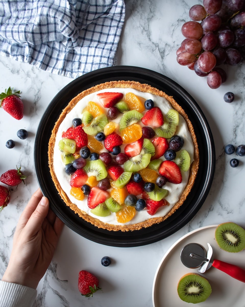 A round fruit pizza with three layers sits on a black tray on a white marbled surface. The bottom layer is a thick, golden-brown crust with a slightly rough texture. The middle layer is a smooth, creamy white spread covering the crust evenly. The top layer has colorful fruits scattered all over, including bright green kiwi slices, red grape halves, strawberry quarters with red and white tones, small dark blue blueberries, and shiny orange mandarin segments. Around the tray are whole fruits, like a bunch of purple grapes, halved kiwi, and whole strawberries. A white plate with a pizza cutter handle in soft red rests nearby on the white marbled surface, and a woman's hand holds the edge of the black tray. There is a blue and white checkered cloth casually placed in the top corner. Photo taken with an iphone --ar 4:5 --v 7