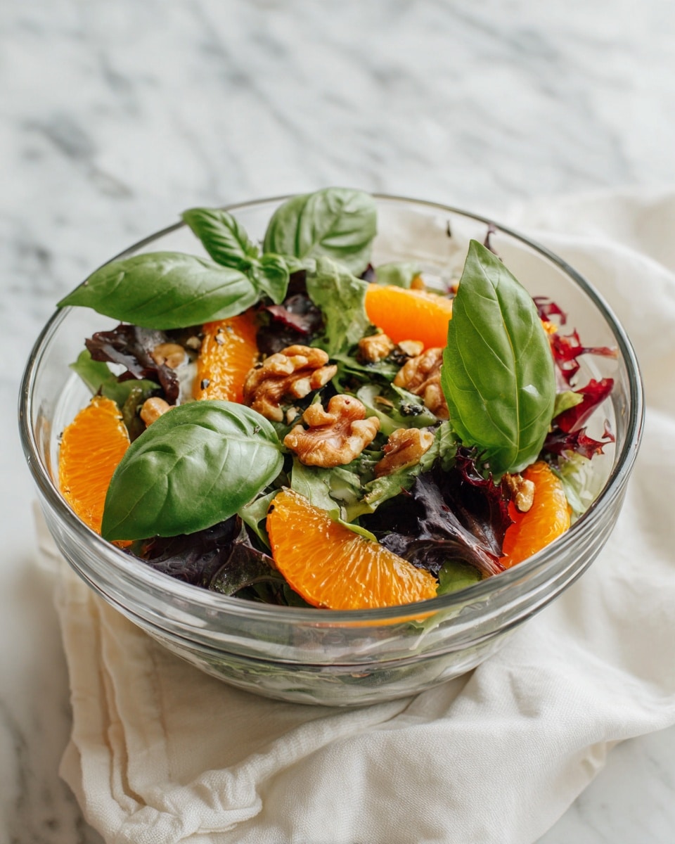 A clear glass bowl holds a colorful salad with two layers: the bottom layer is mixed green leaves with different shapes and dark green colors, and the top layer has bright orange and red round slices of citrus fruits. Small pieces of light brown walnuts are spread over the salad, along with a few whole deep green basil leaves placed on top. The bowl sits on a white marbled surface with a white cloth nearby. Photo taken with an iphone --ar 4:5 --v 7