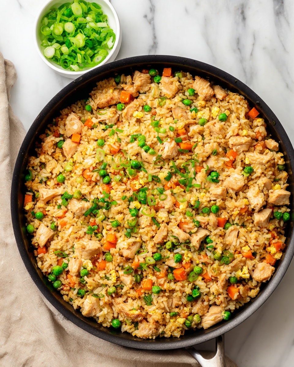 A close-up view of a large black pan filled with fried rice. The dish has several layers: small pieces of light brown cooked chicken scattered evenly throughout, bright orange cubes of cooked carrots, green peas, and chopped green onions sprinkled on top, adding a fresh green color. The rice is golden brown, mixed with the other ingredients, and looks soft and well-cooked. The pan is on a white marbled surface, with a small white bowl filled with sliced green onions positioned in the upper left corner. There is a beige cloth partially under the pan on the left side. The photo taken with an iphone --ar 4:5 --v 7