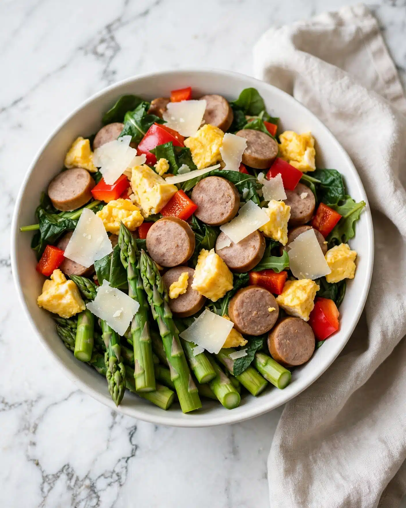 Sheet Pan Sausage and Tortellini Recipe 5 square image This image shows a white bowl filled with a colorful meal featuring several layers. At the bottom, there are bright green asparagus pieces standing out among other ingredients. On top of this green layer, there are round slices of light brown sausage and chunks of yellow egg. Mixed among these are vibrant red bell pepper pieces and some darker green leafy vegetables. Scattered over everything are thin slices of light-colored cheese, adding texture. The bowl sits on a white marbled surface with a soft cloth nearby. The light in the photo highlights the fresh and varied textures of the dish, making it look tasty and fresh. photo taken with an iphone --ar 4:5 --v 7