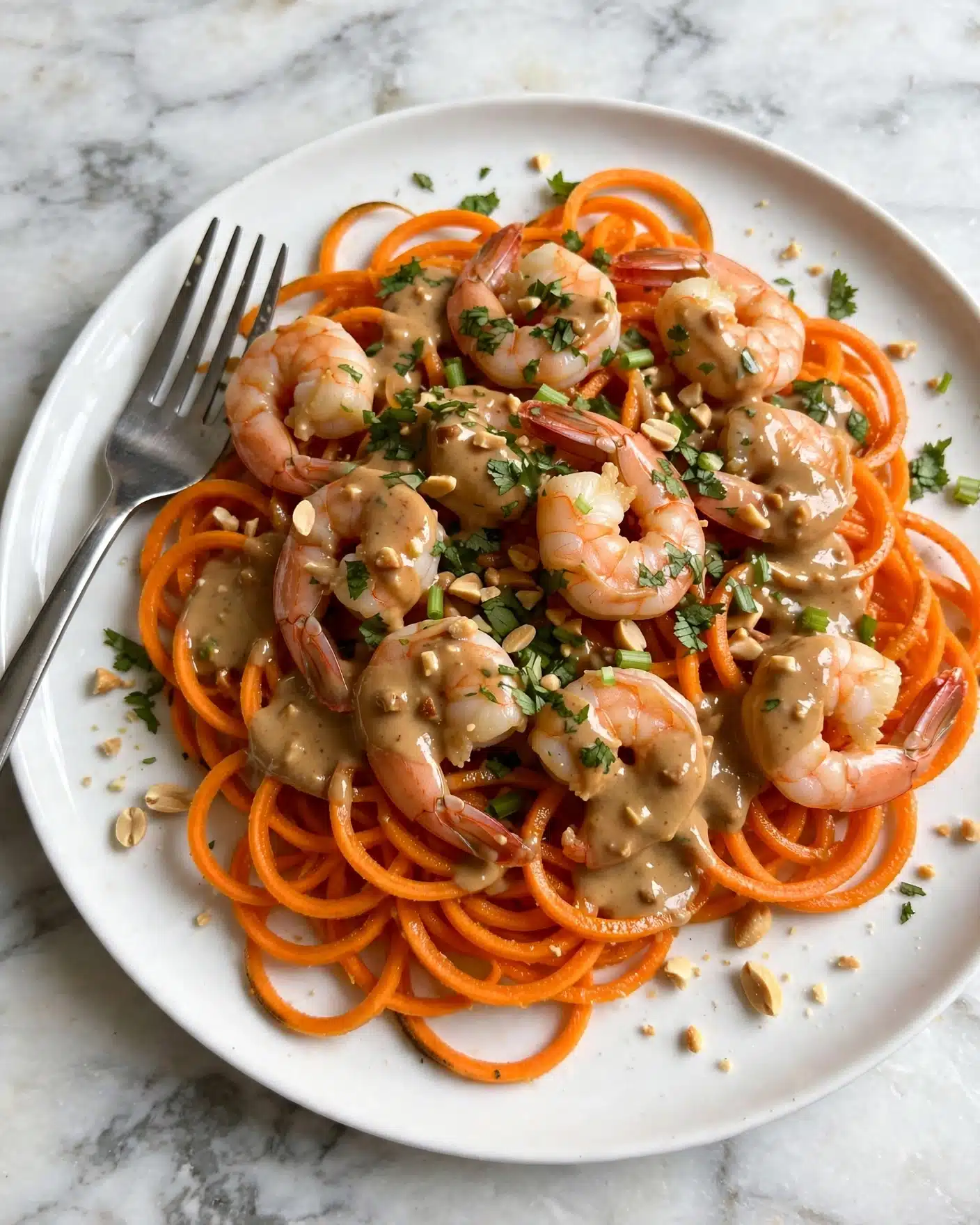 Quick Coconut Shrimp Noodles Recipe 5 square image A white plate holds a dish with three main layers: the bottom layer is bright orange spiralized sweet potato noodles with a slightly shiny texture, spread evenly across the plate. On top are cooked shrimp with a light pink color, placed around and in the middle of the noodles. Covering both is a creamy light brown sauce with visible small bits, drizzled over the shrimp and noodles. The dish is sprinkled with chopped green herbs and scattered chopped peanuts, some resting on the food and some on the plate edge. A silver fork sits on the left edge of the plate. The scene is set on a white marbled surface. photo taken with an iphone --ar 4:5 --v 7